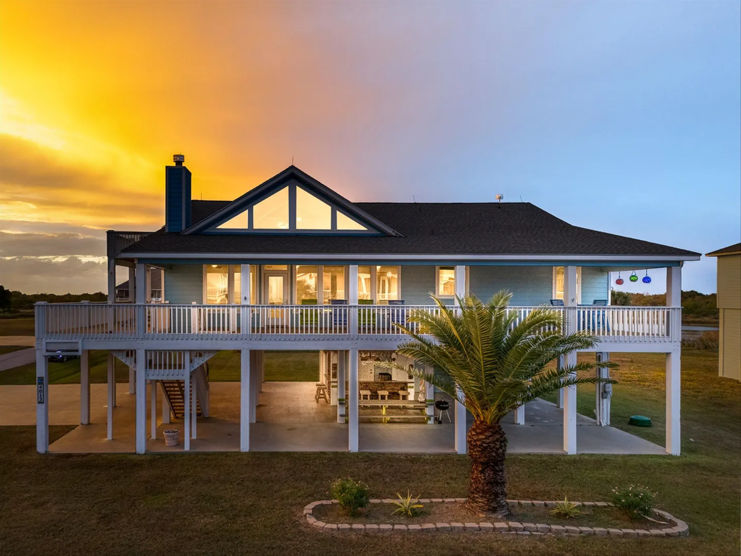 Exterior dusk view of a stilted beach house with illuminated interior lights, an upper wrap-around deck, and ground-level patio against a colorful sunset sky.