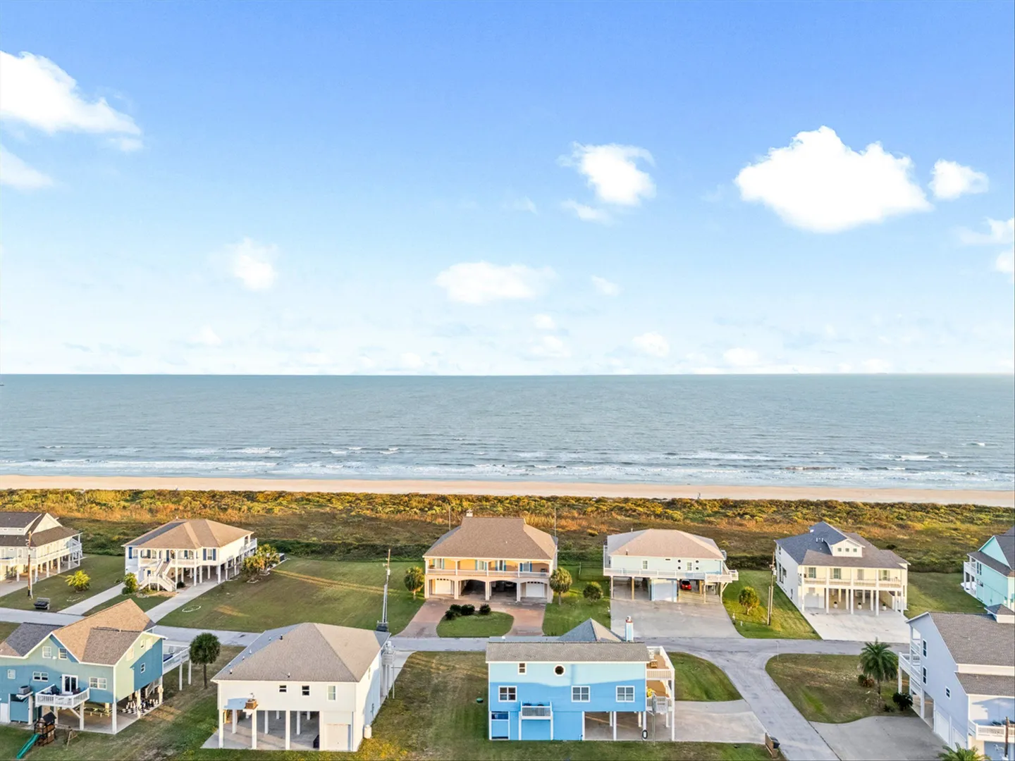 Aerial view of a coastal neighborhood with stilt homes near the sandy beach and ocean under a blue sky.