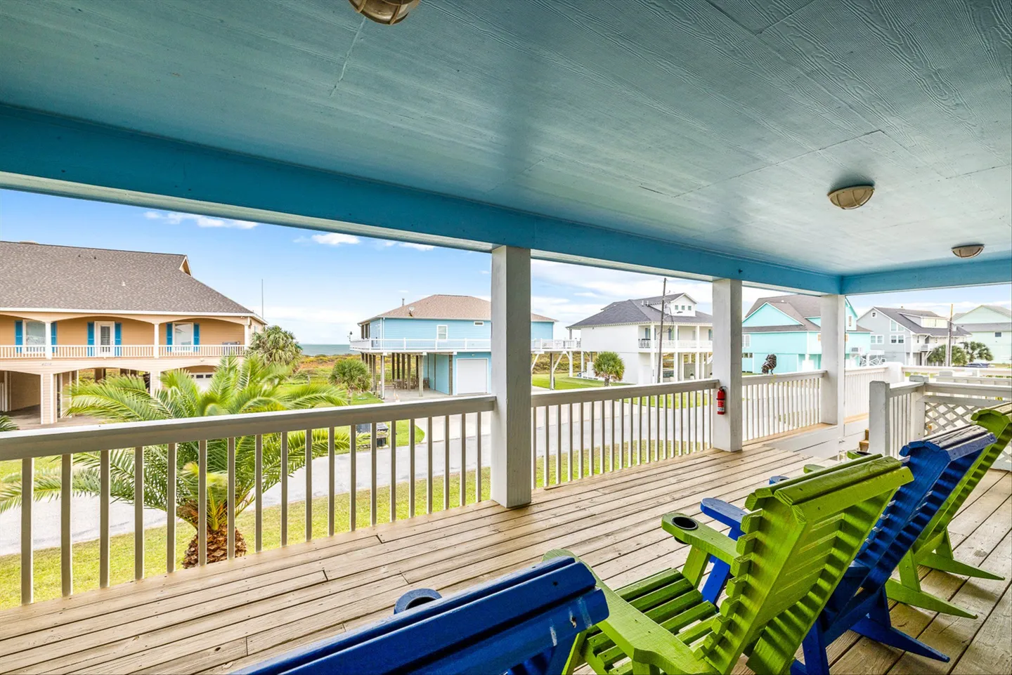 Covered deck with wooden flooring, colorful chairs, and a white railing overlooking a neighborhood of stilt houses under a blue sky.