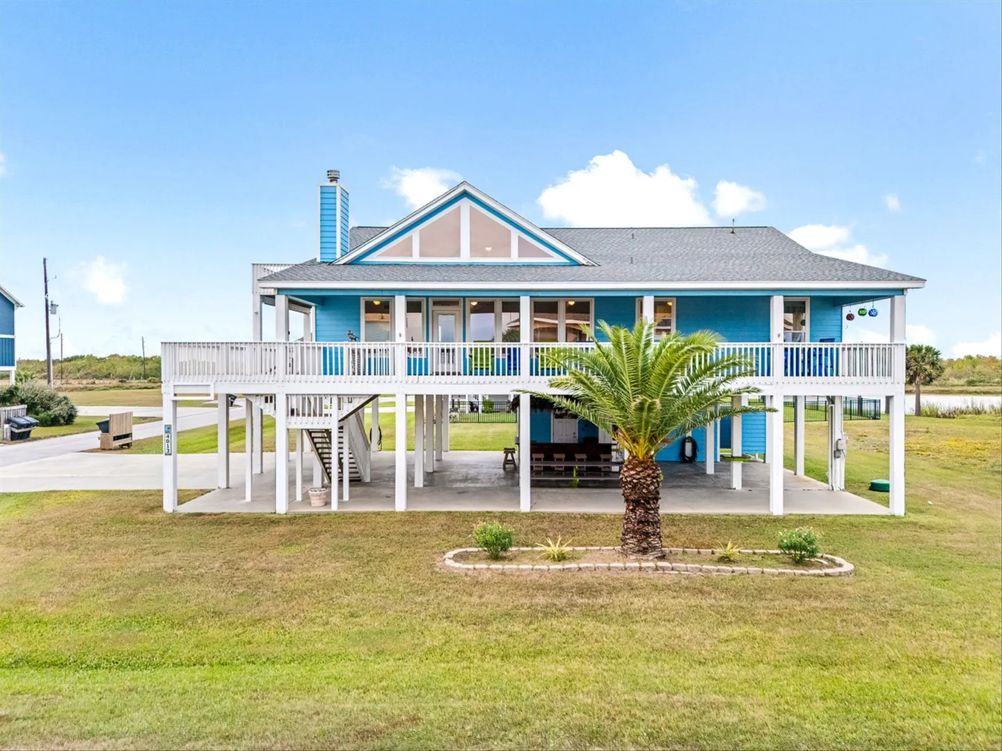 Exterior view of an elevated bright blue beach house on stilts with an expansive wrap-around deck and covered ground-level space.