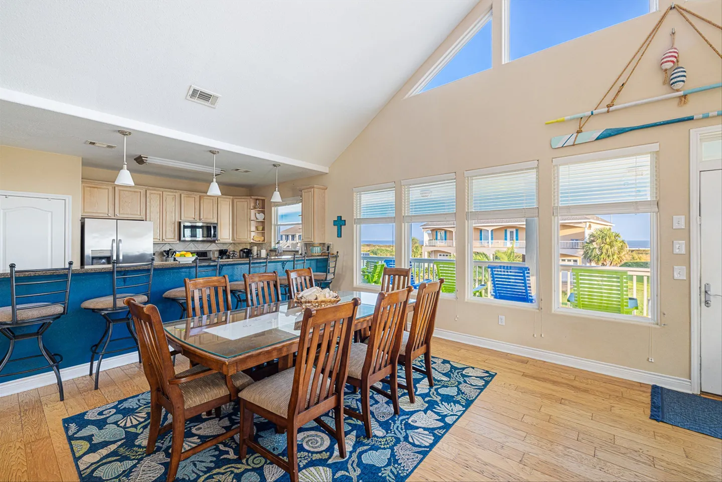 Dining area with a large wooden table, patterned blue rug, and adjacent kitchen with a breakfast bar under vaulted ceilings.