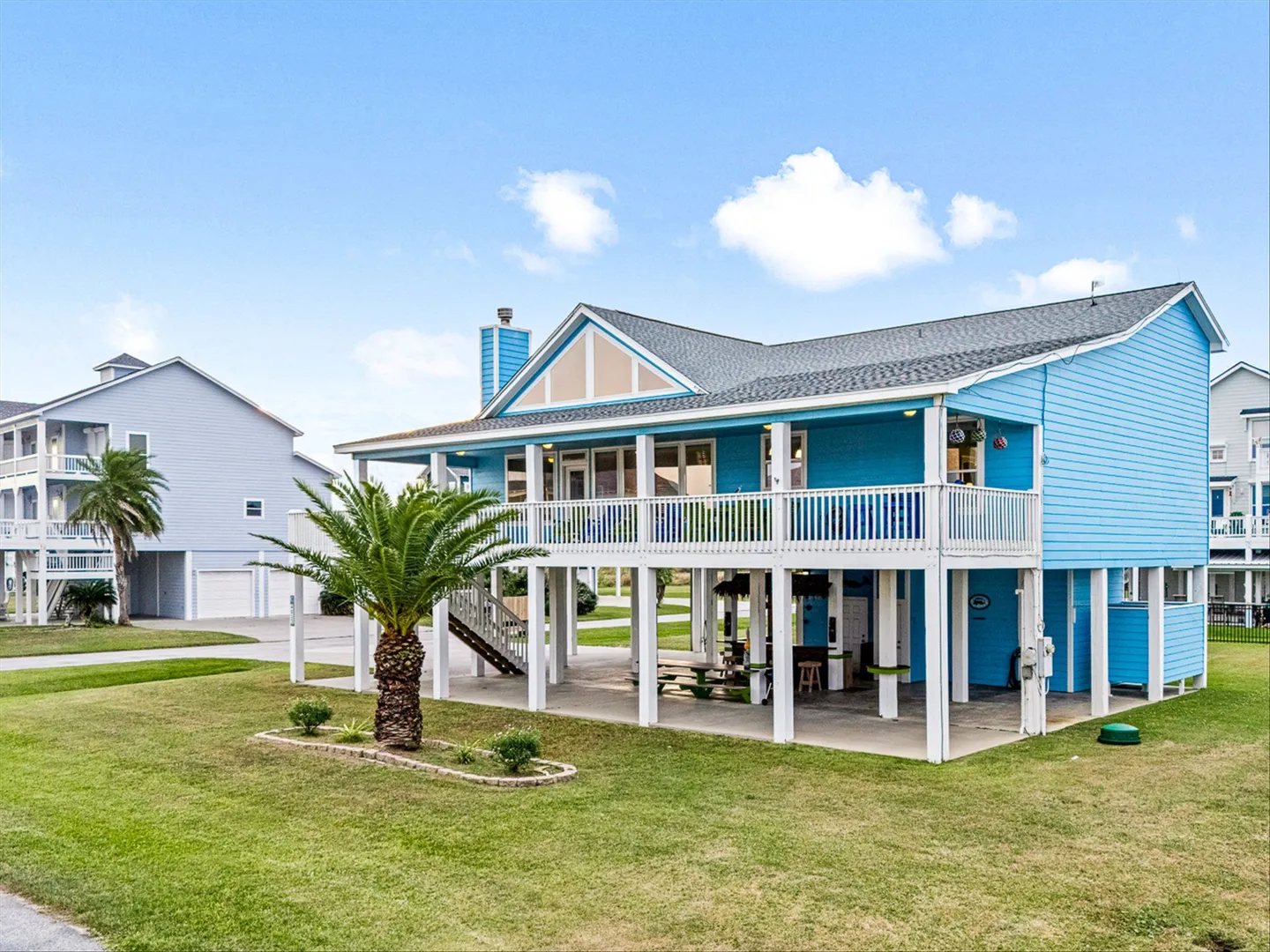 Exterior of a bright blue stilted beach house with a large upper deck and covered ground-level patio space.