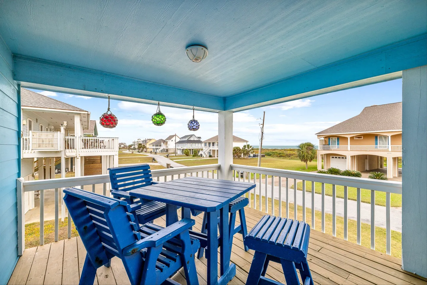 Covered deck with a light blue ceiling, wood plank flooring, and a blue dining table set overlooking a coastal neighborhood.