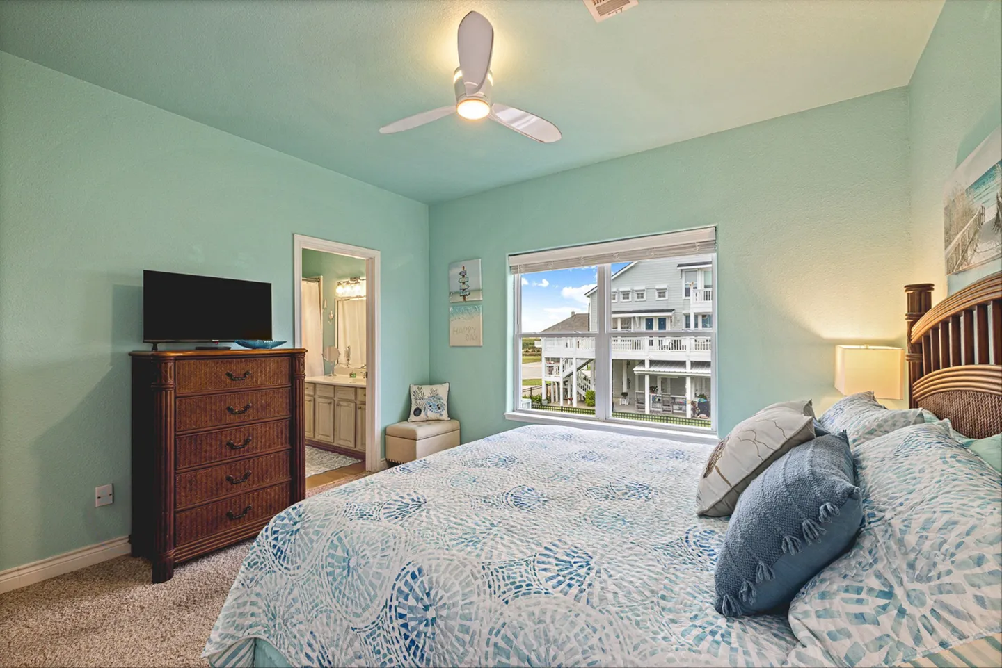 Bedroom with light green walls, a patterned bedspread, ceiling fan, dresser with TV, and an ensuite bathroom doorway.