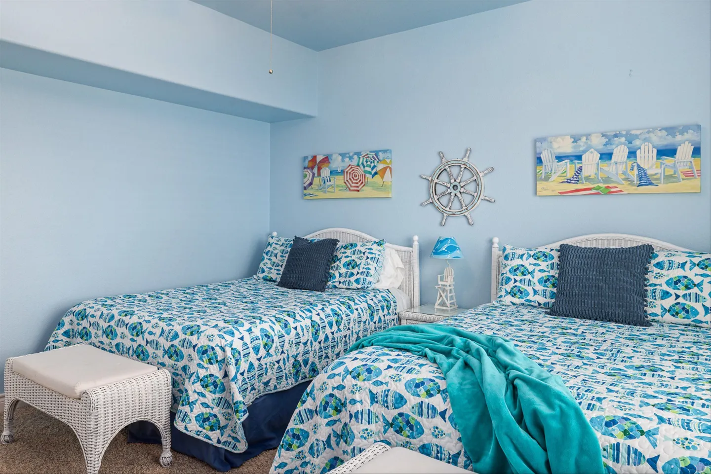 Bedroom with two beds featuring matching blue fish-patterned quilts, white wicker headboards, and light blue walls decorated with coastal art.