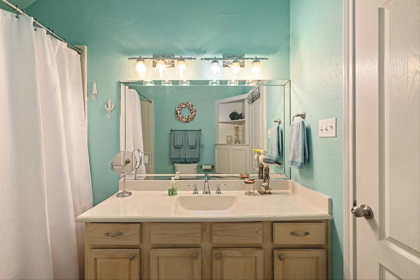 Bathroom with turquoise walls, a light wood vanity, large mirror, vanity lights, and a white shower curtain.