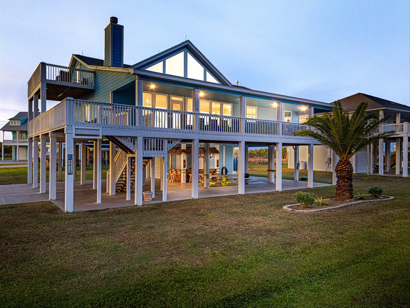 Exterior of an elevated stilt beach house with a wraparound deck, lit windows at dusk, and a sheltered patio area underneath.