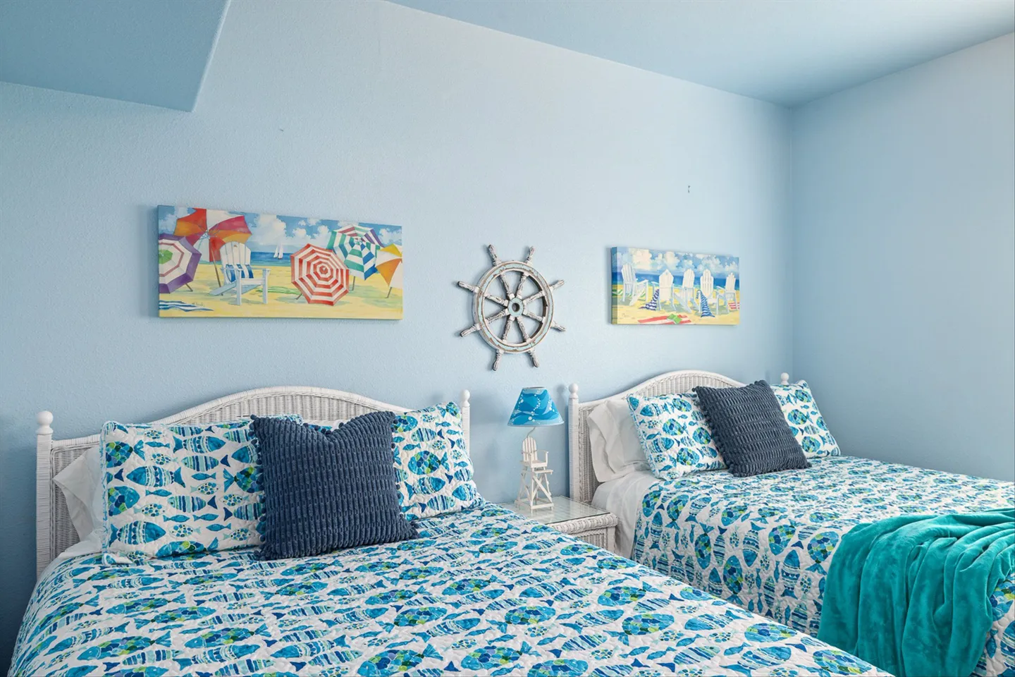 Bedroom with two beds featuring white wicker headboards, fish-patterned quilts, and coastal decor on light blue walls.