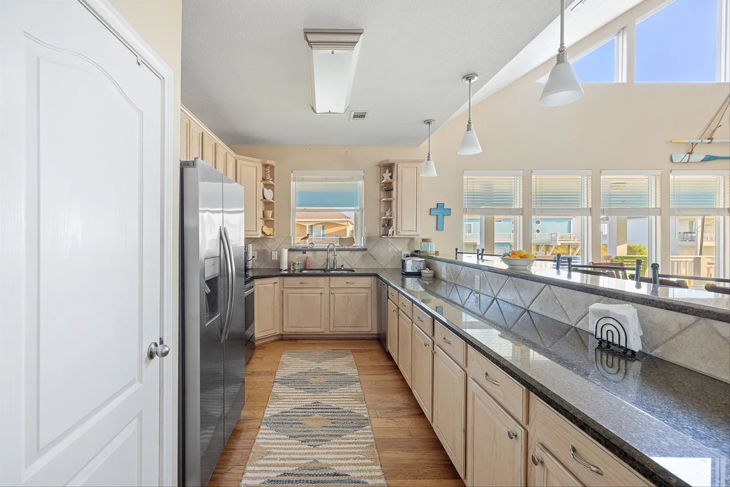 Bright kitchen featuring light wood cabinetry, dark speckled countertops, a patterned runner rug, and stainless steel refrigerator.