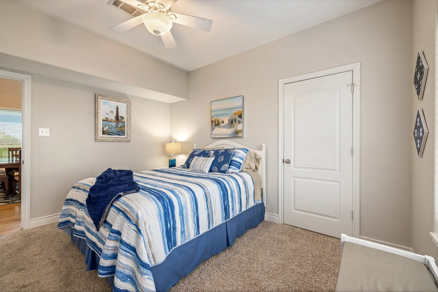 Bedroom with carpet, a white headboard bed dressed in blue and white striped bedding, and coastal artwork on neutral walls.