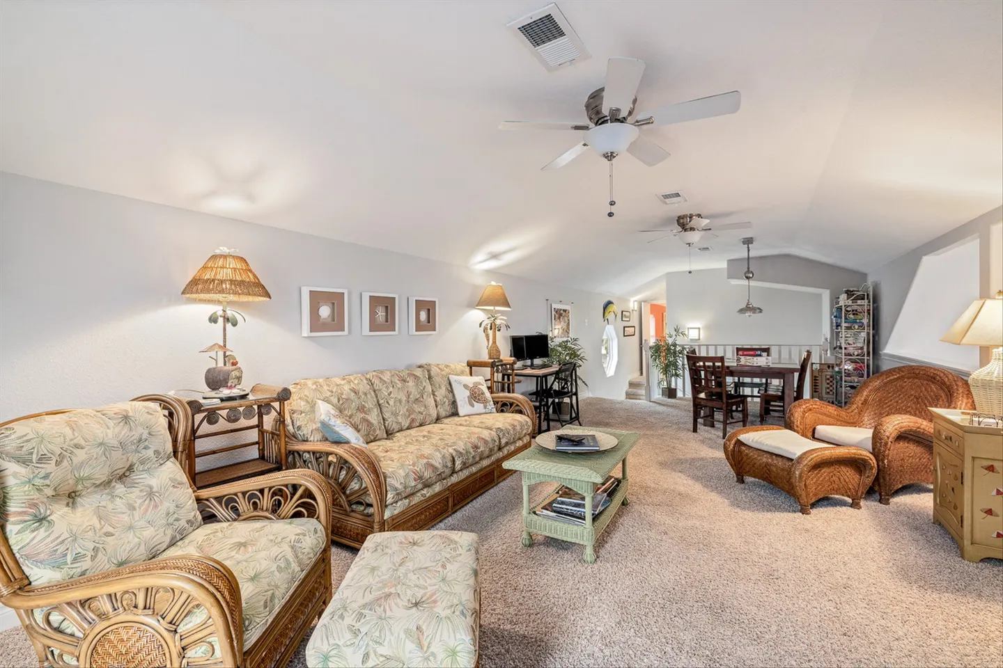 Open-concept living room with wicker and rattan furniture, patterned cushions, carpeted floors, and a dining area in the background.