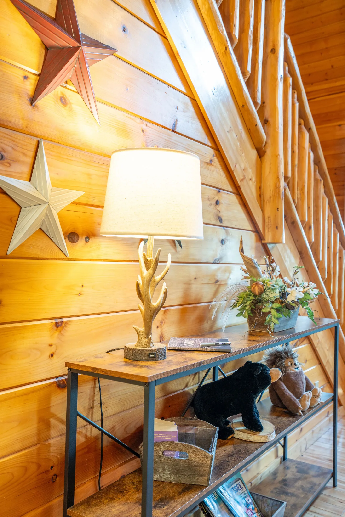 A rustic console table beneath a wooden log staircase with an antler lamp and star wall decor.