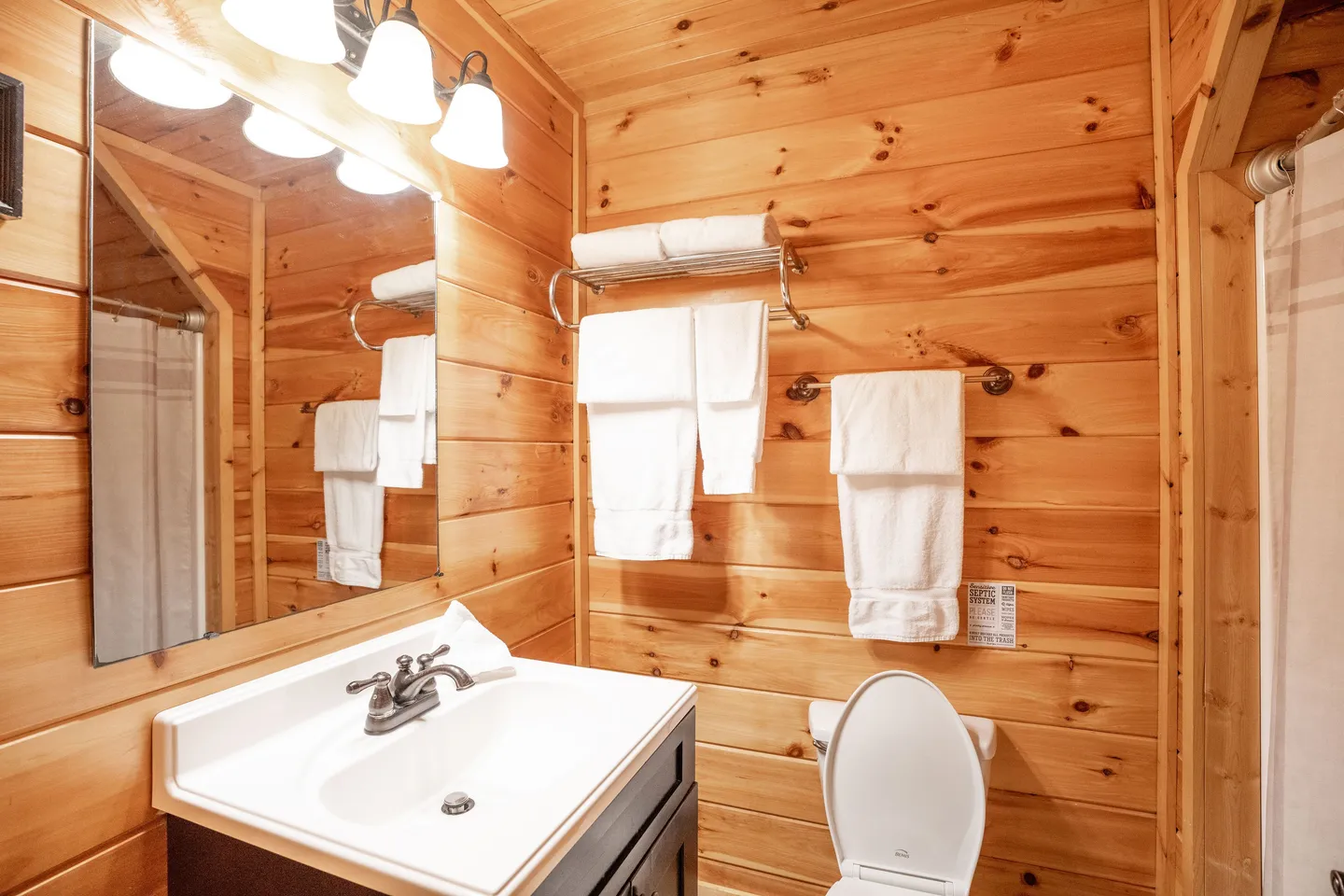 Bathroom with wood-paneled walls, a vanity with a white sink and mirror, a toilet, and towel racks with folded white towels.