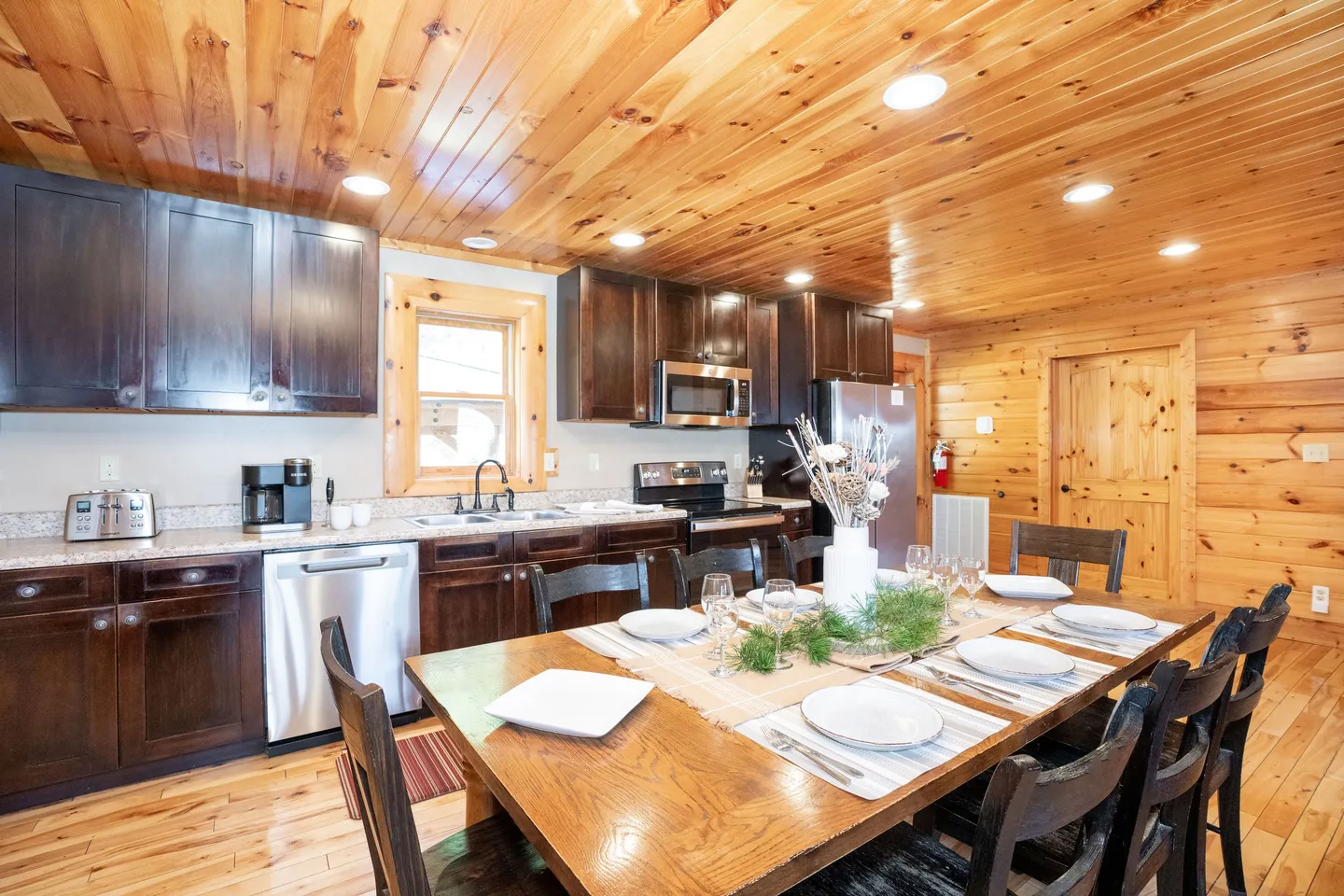 Dining area with a large wooden table set for a meal adjacent to an open kitchen with dark cabinets and stainless steel appliances.