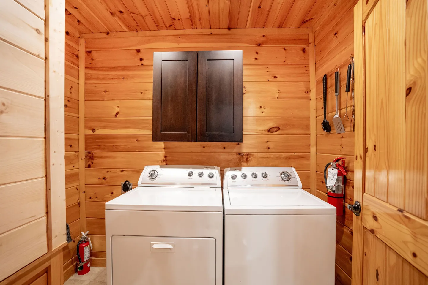 A laundry alcove with wood-paneled walls, featuring a white washer and dryer beneath a dark wooden upper cabinet.
