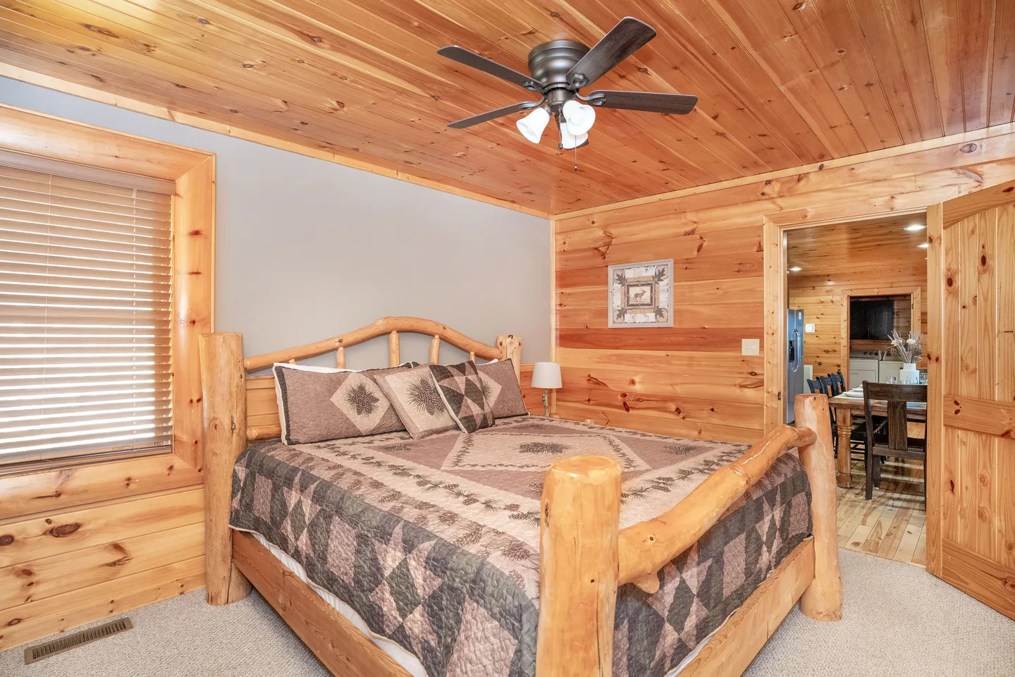 Bedroom with a log-frame bed, wood-paneled walls, ceiling fan, and view into the adjacent dining area.
