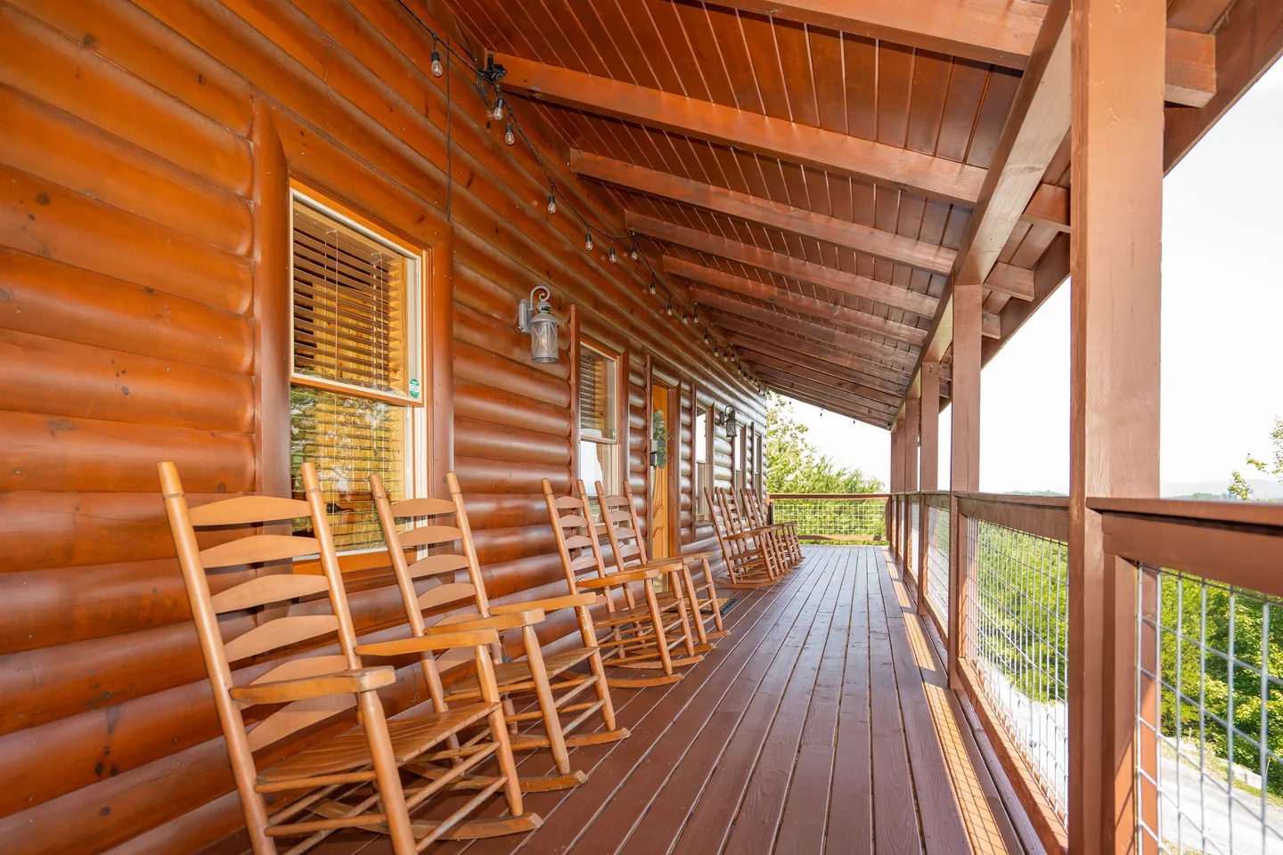 A covered wooden deck of a log home featuring a row of wooden rocking chairs overlooking trees.