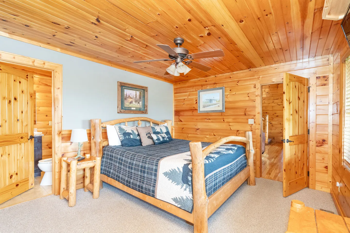 Rustic bedroom with log bed frame, wood-paneled walls and ceiling, carpeted floor, ceiling fan, and open doors to an en suite bathroom and hallway.