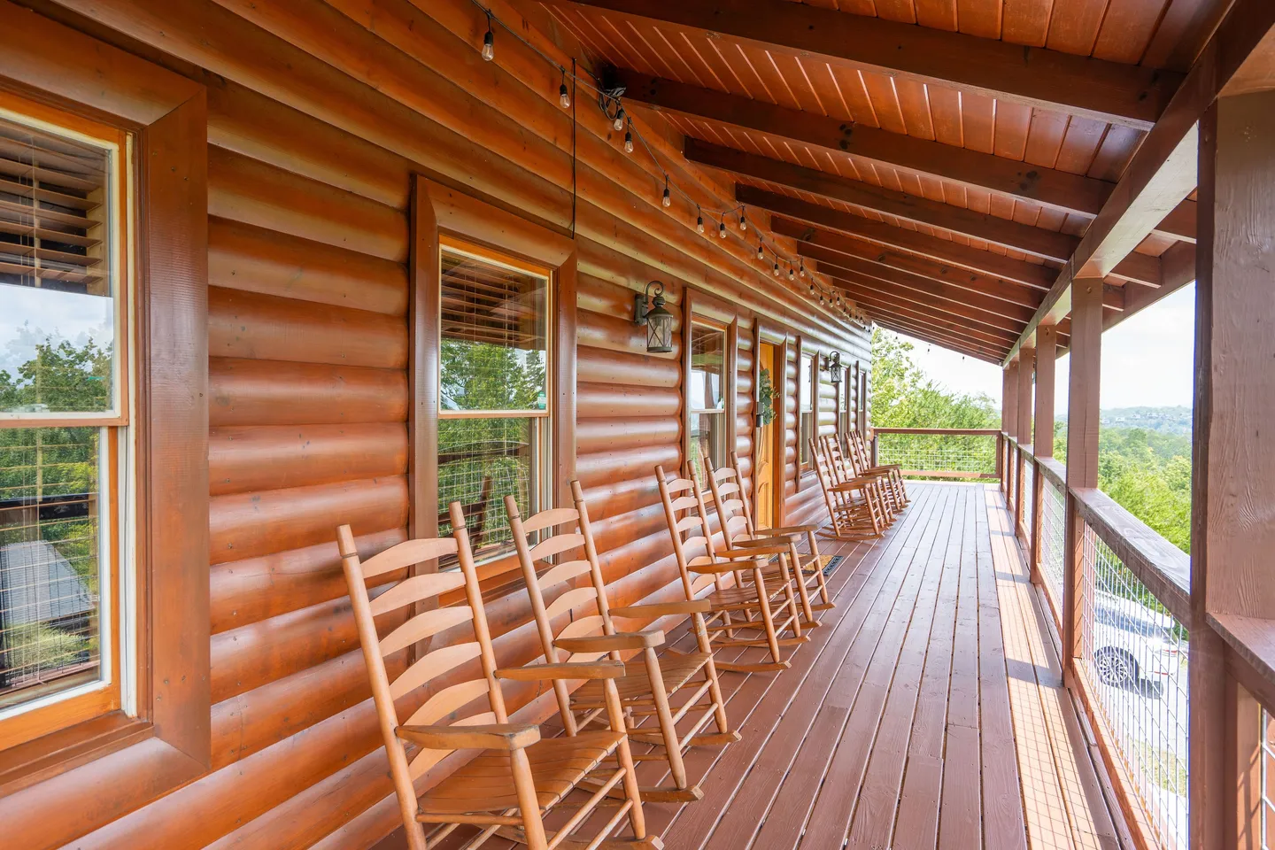 A covered wooden deck of a log cabin lined with rocking chairs overlooking outdoor scenery.