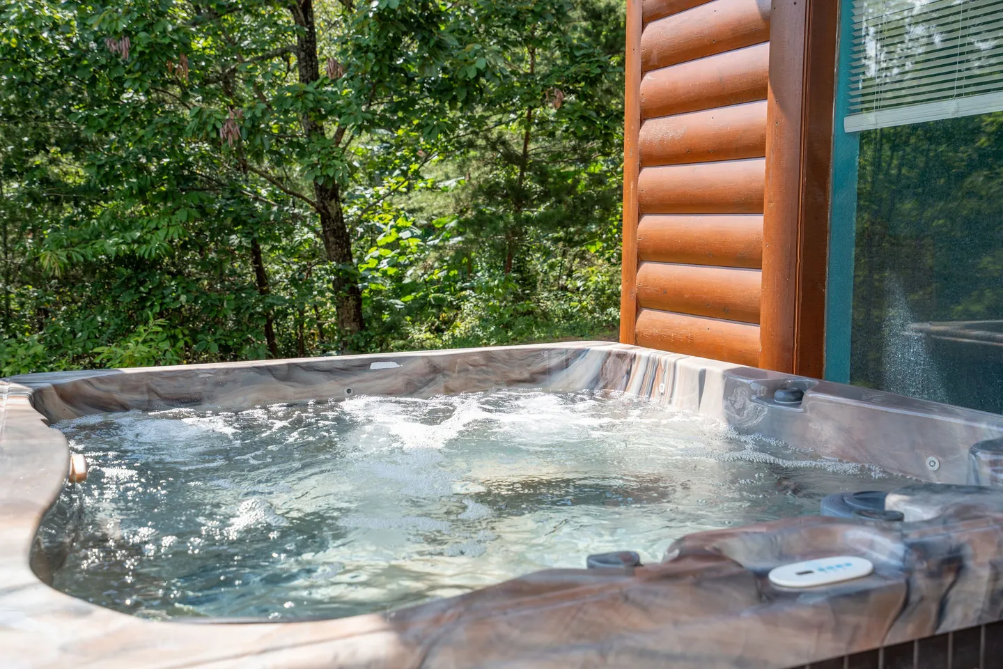 Close-up of a bubbling outdoor hot tub on a deck next to a log cabin exterior wall, surrounded by green trees.