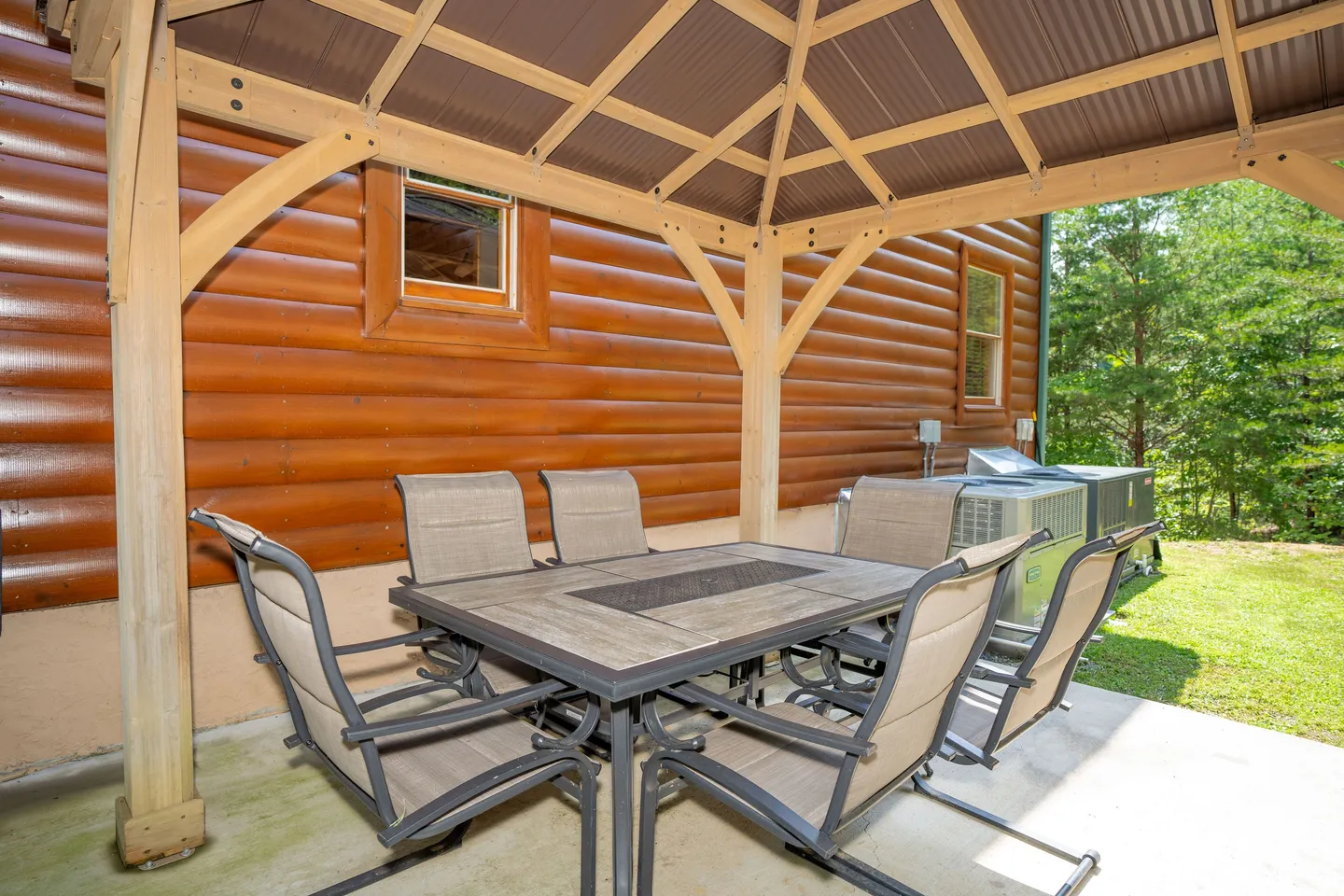 Outdoor patio dining table with chairs under a wooden pavilion next to a log cabin exterior wall.