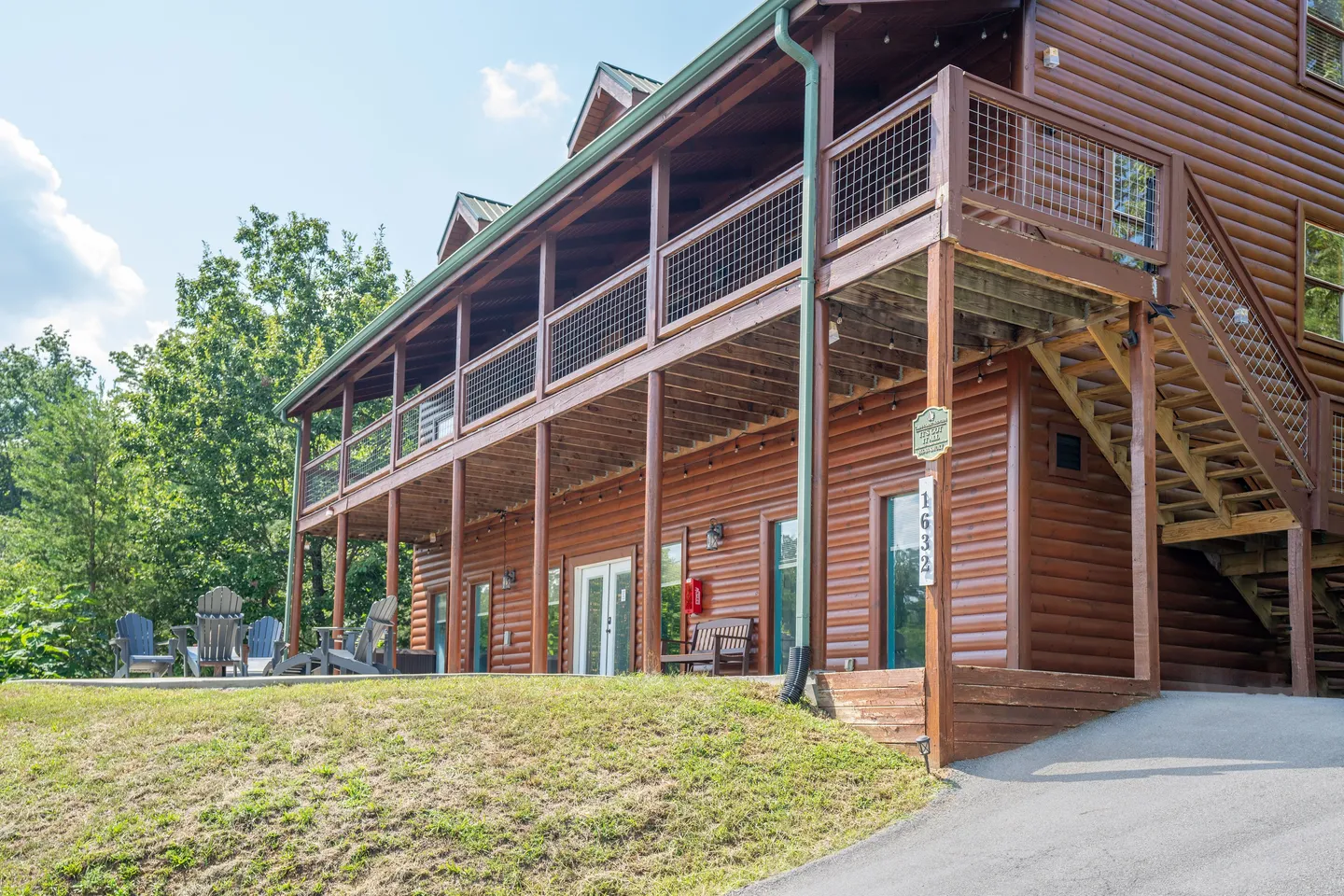 Exterior of a multi-story log cabin showing a two-level covered deck, outdoor seating, and a paved driveway under a sunny sky.