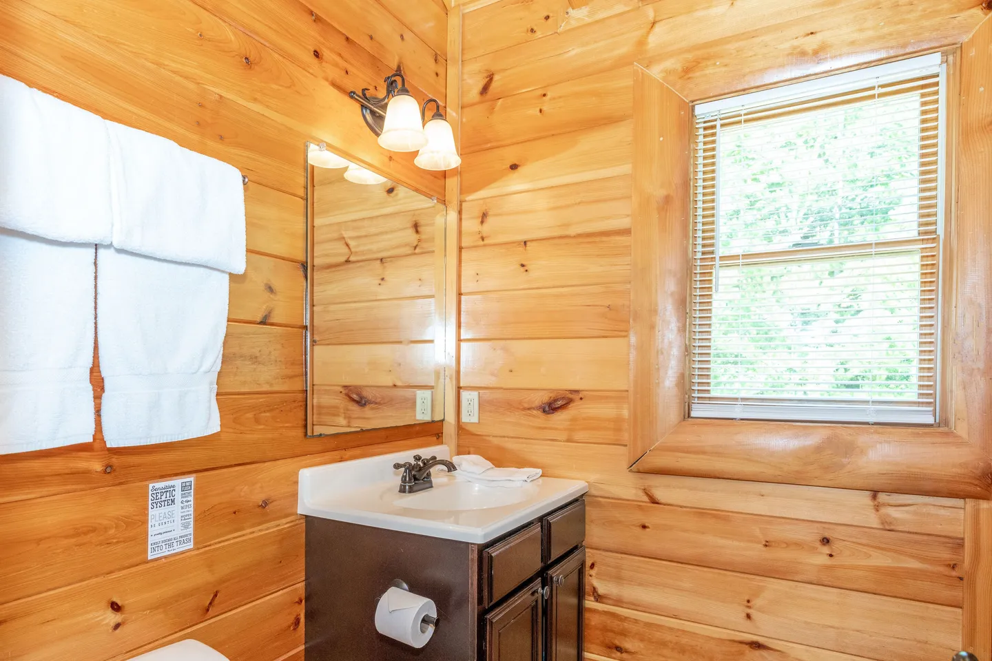 Bathroom with wood-paneled walls, vanity sink, mirror, wall sconce, and a window with blinds.