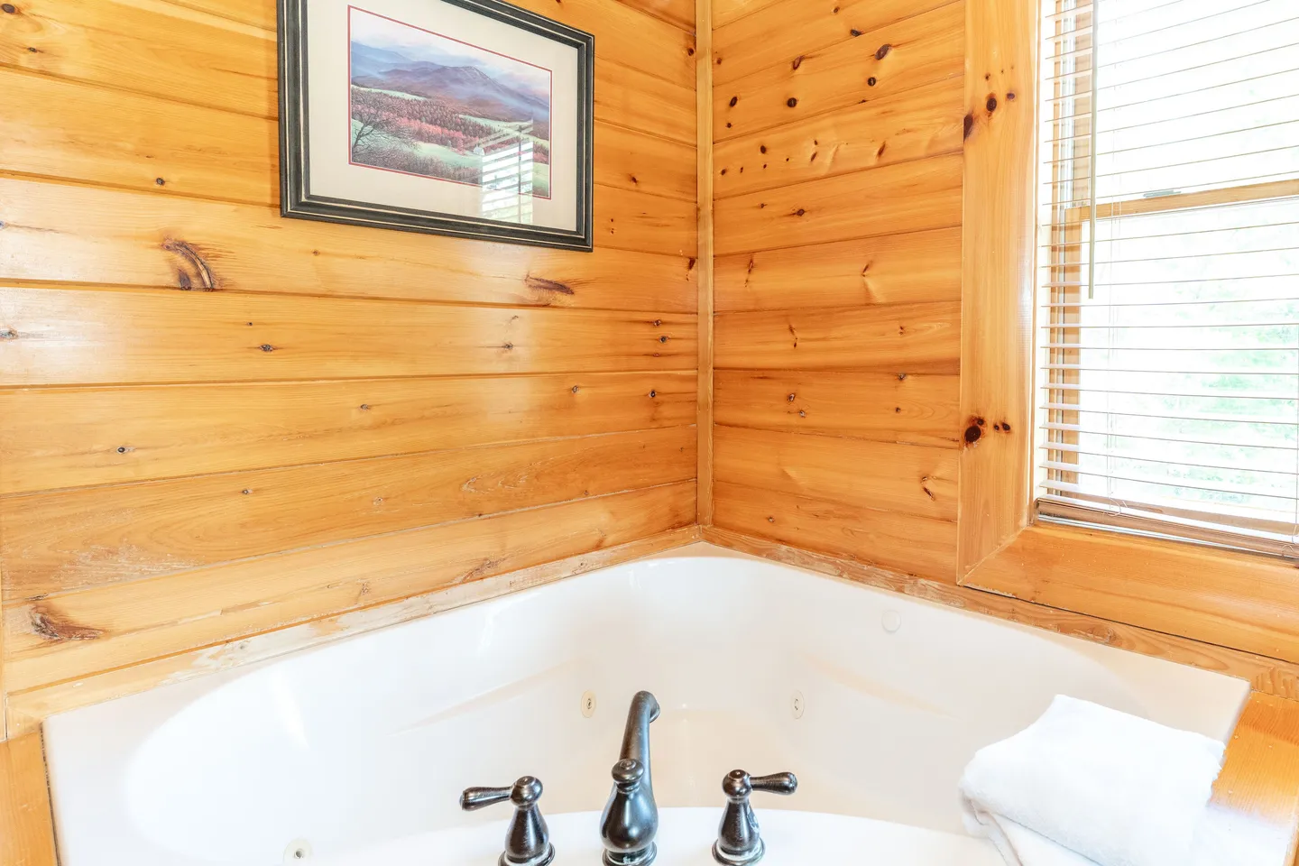 Corner jetted tub set against knotty wood-paneled walls beneath a window with blinds.