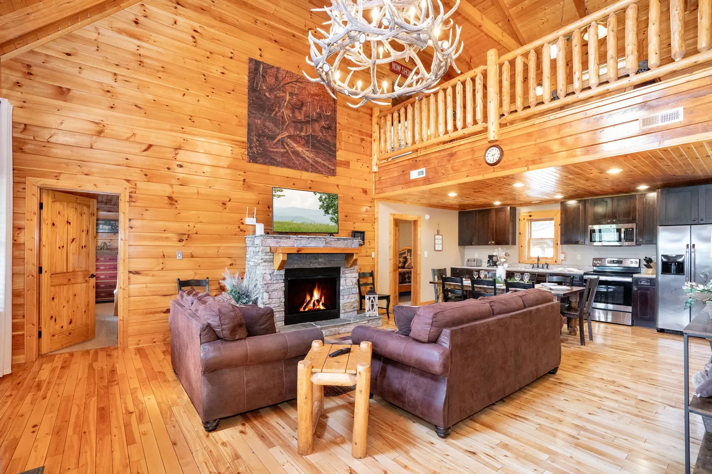 Open-concept log cabin living room with high vaulted wood ceilings, an antler chandelier, a stone fireplace, brown sofas, and a view into the kitchen.