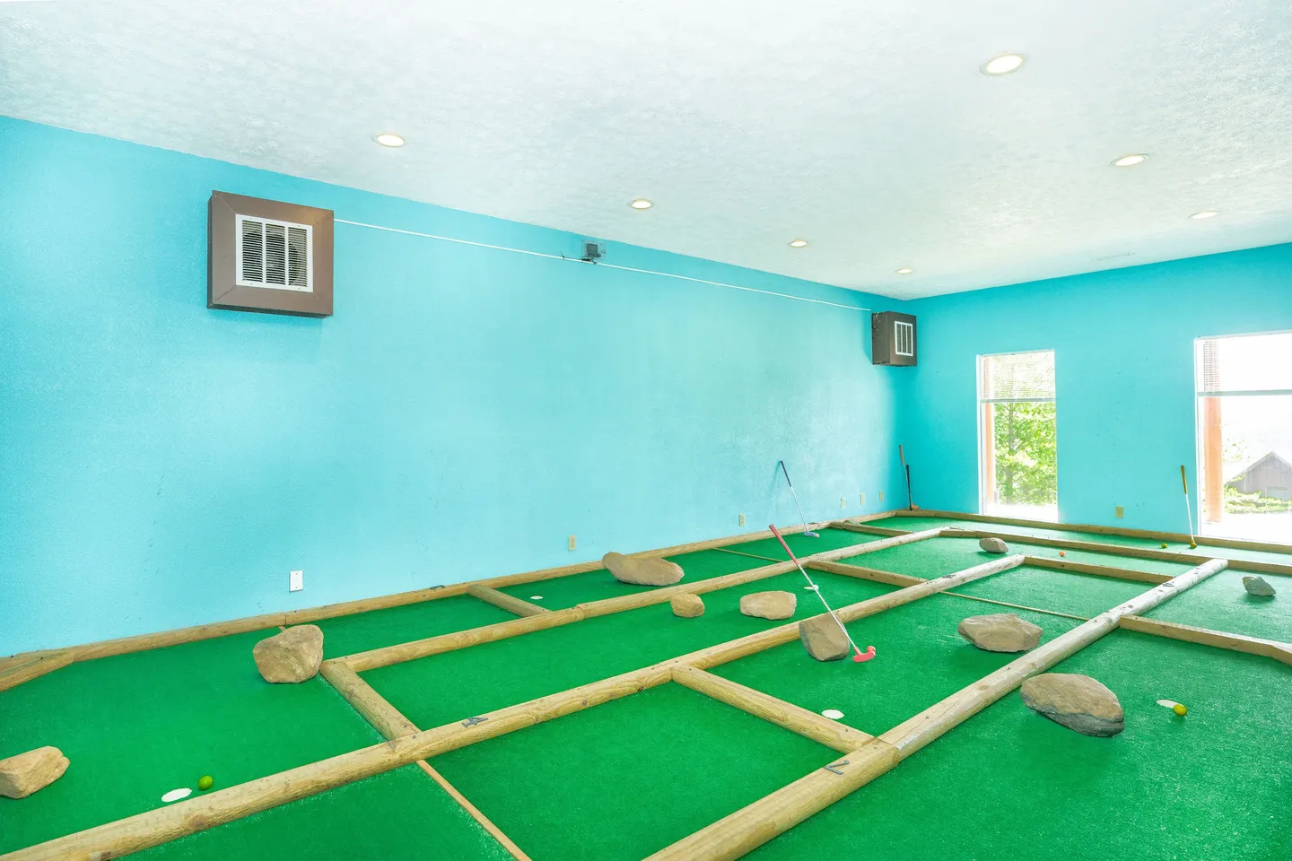 Indoor miniature golf room with bright turquoise walls, artificial turf, wooden borders, rocks as obstacles, and putters.