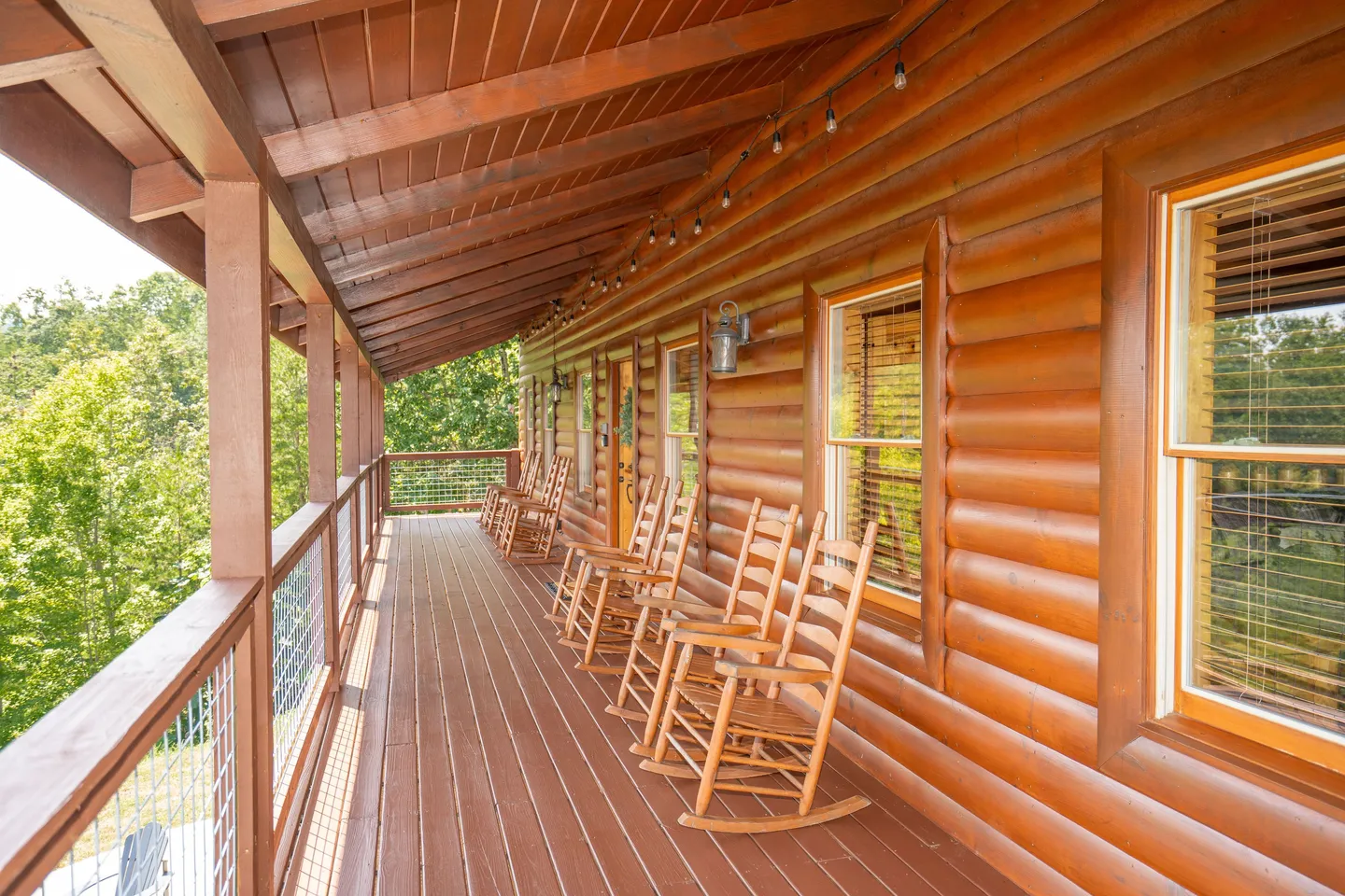 A covered wooden deck of a log home featuring a row of rocking chairs overlooking trees.
