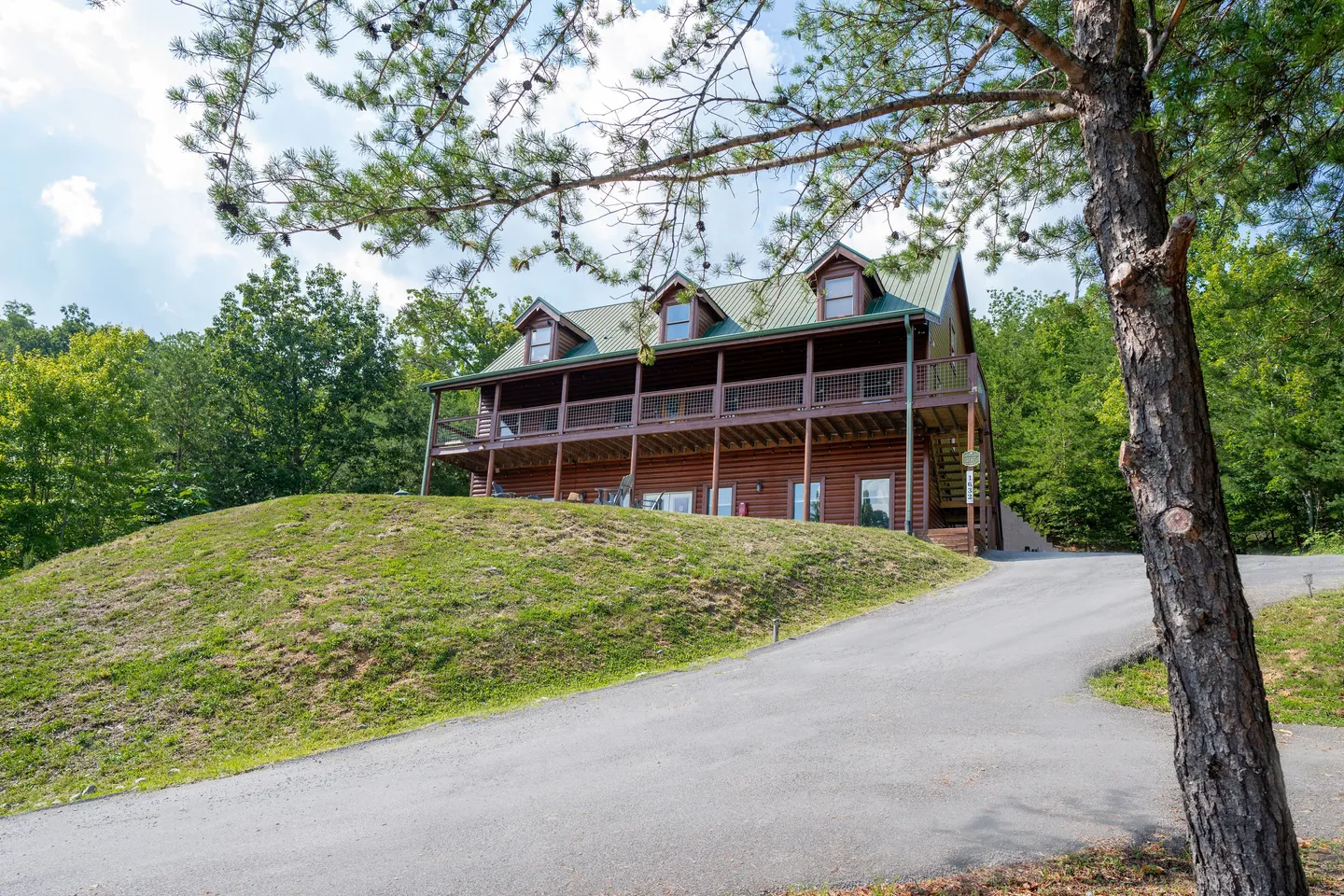 Exterior view of a multi-story log cabin on a grassy hill with a paved driveway leading up to it under a blue sky.