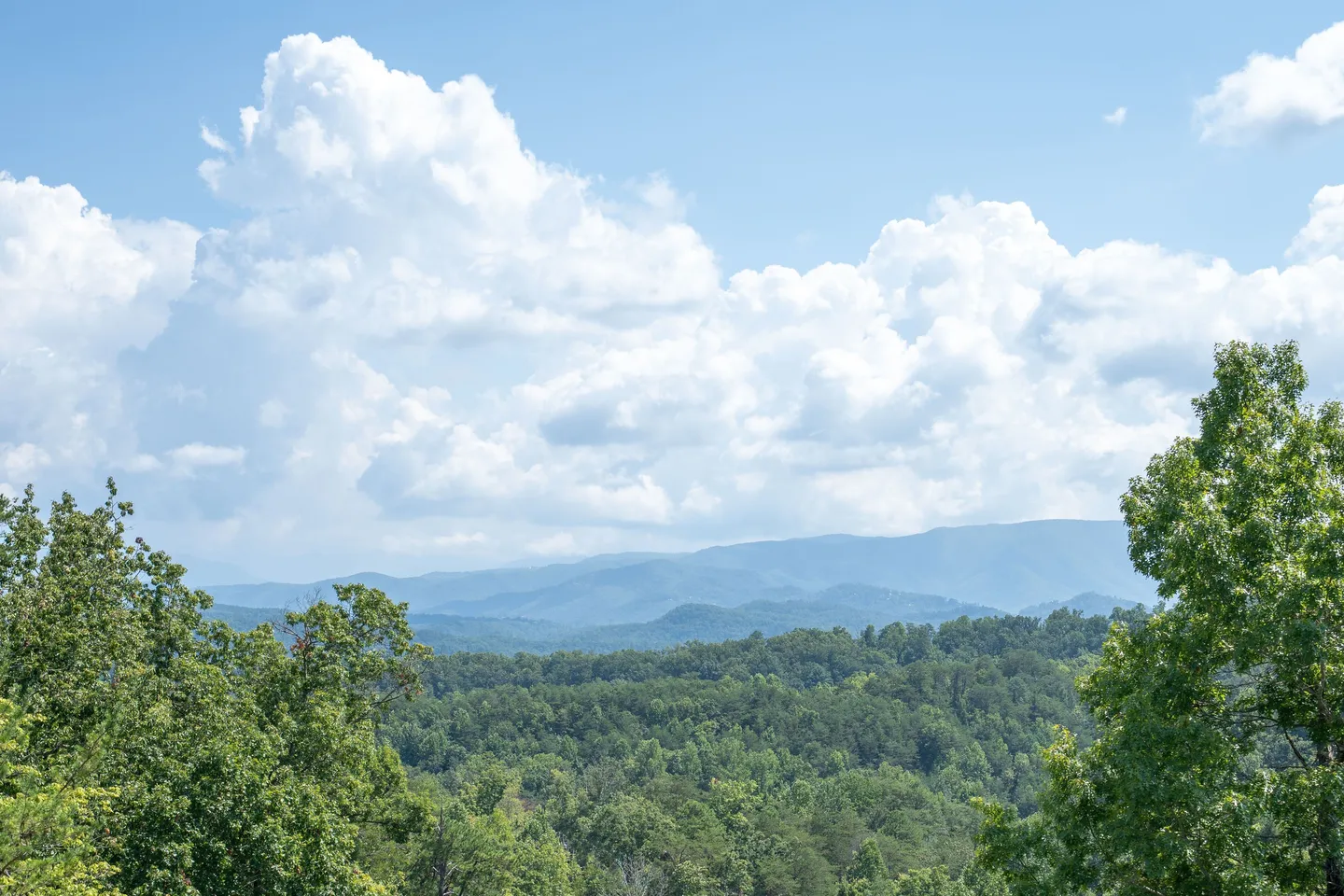 Elevated scenic view of green forested hills stretching out toward blue-toned mountain ridges under a partly cloudy sky.