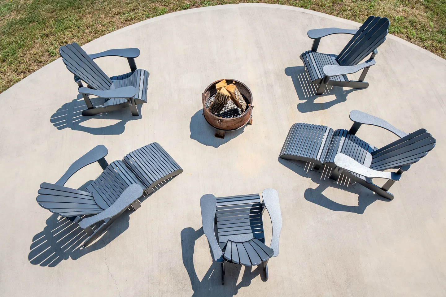 High-angle view of a circular concrete patio with a central fire pit containing firewood, surrounded by dark Adirondack chairs.