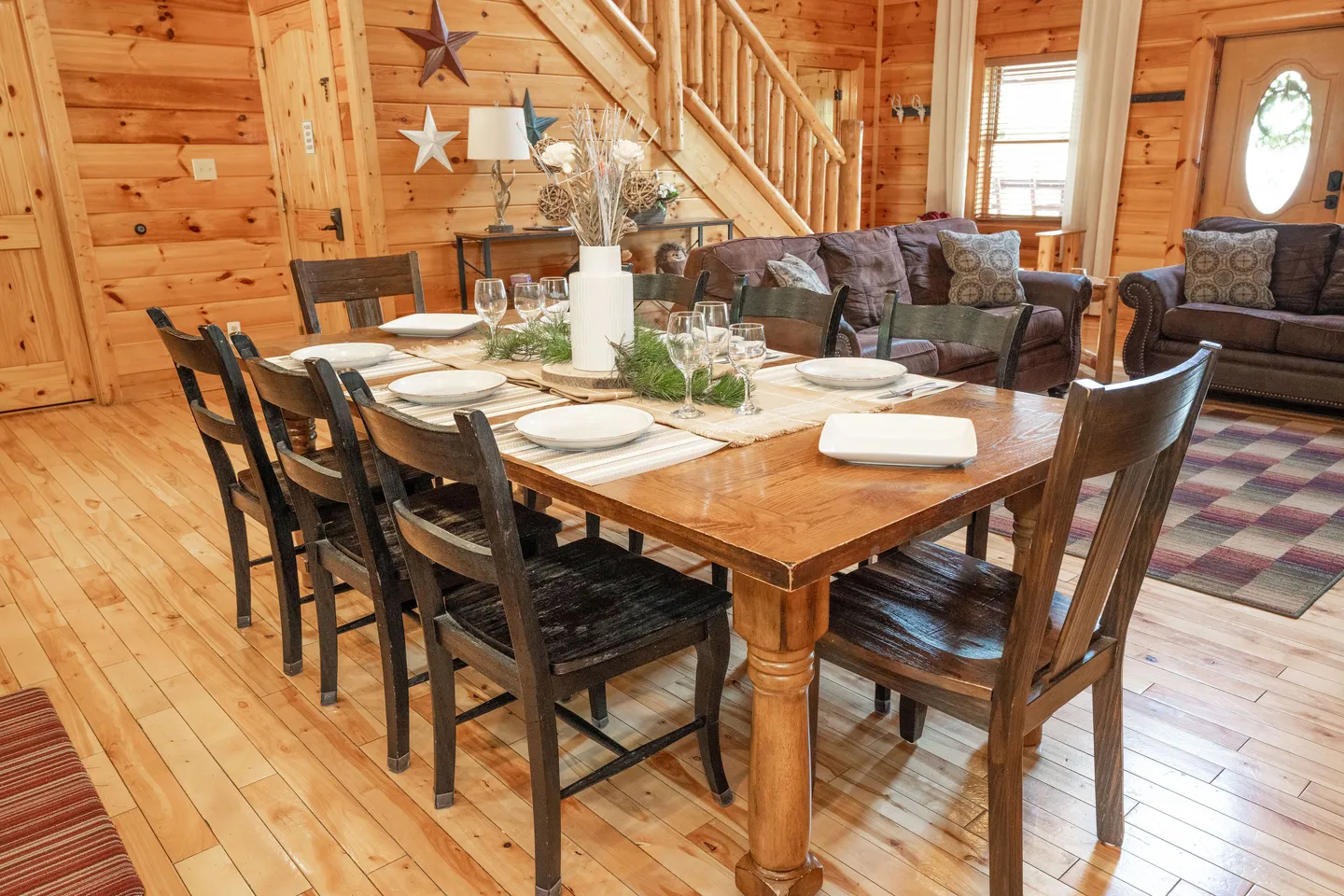 A wooden dining table set with white plates, wine glasses, and a centerpiece, surrounded by dark chairs in a log-walled room.
