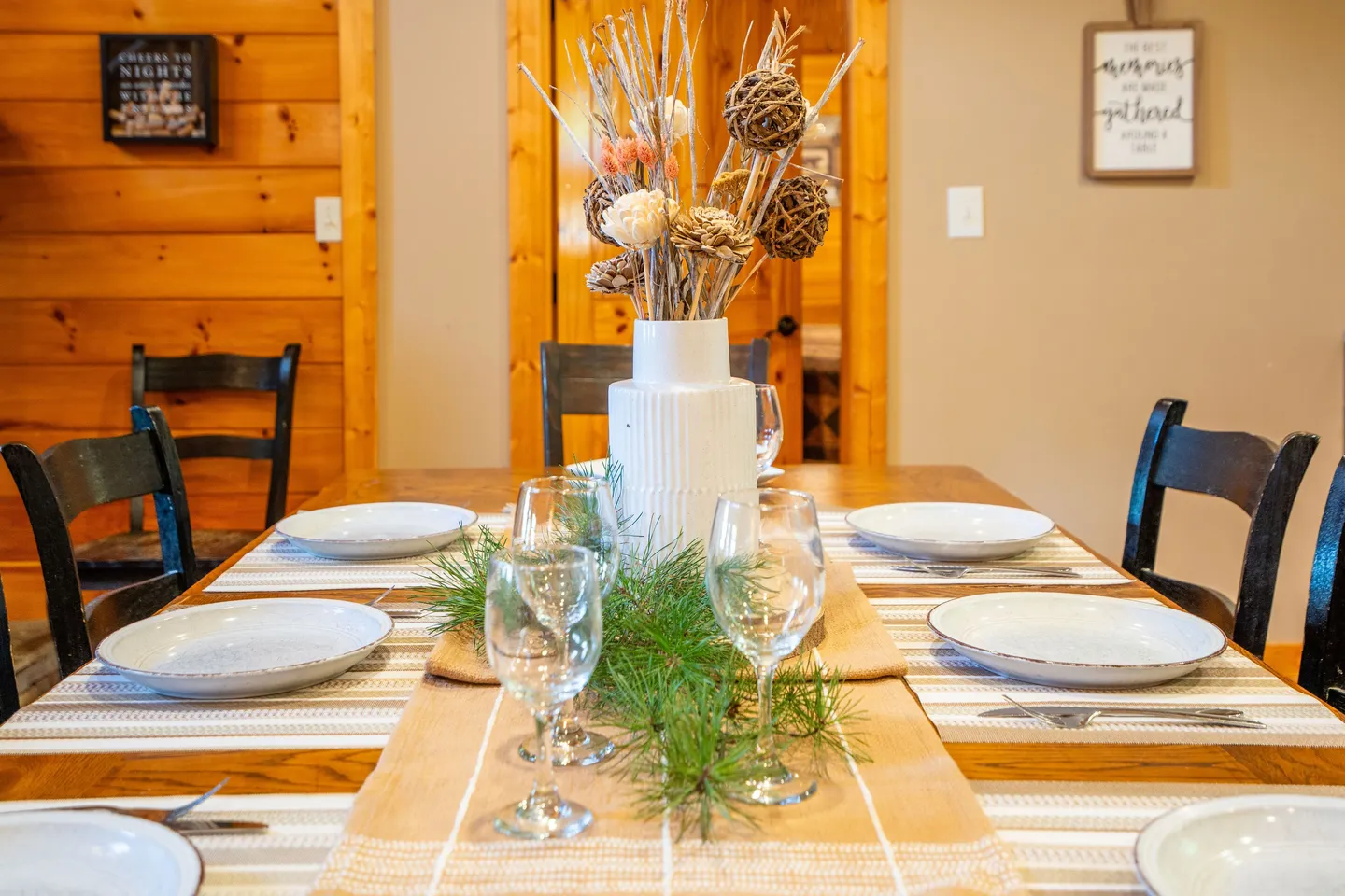 A wooden dining table set with place mats, white plates, wine glasses, and a decorative floral centerpiece.