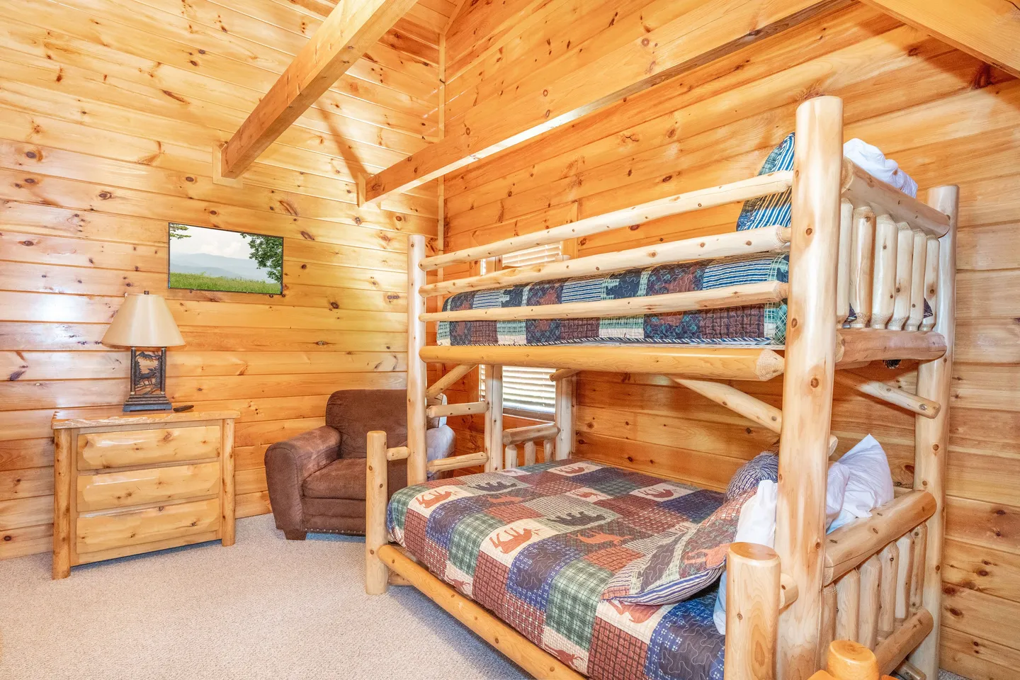 Bedroom with log bunk beds, wood-paneled walls and ceiling, carpeted floor, an armchair, and a small dresser with a lamp.