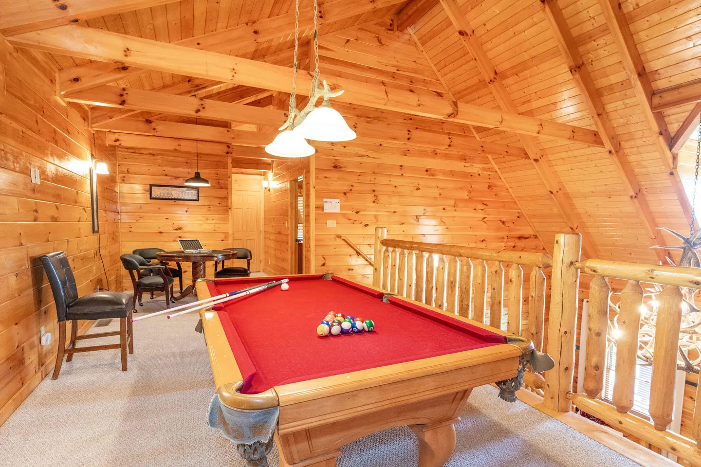 Game room loft in a log cabin featuring a red felt pool table, wood-paneled walls and ceiling, and a card table in the background.