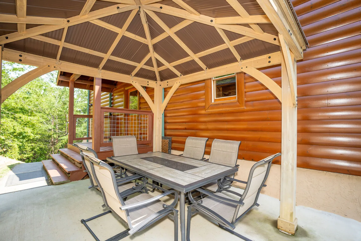 A covered outdoor gazebo next to a log cabin, featuring a dining table and chairs.
