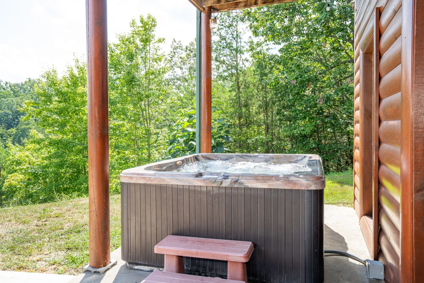 A hot tub with bubbling water on a concrete patio next to a log cabin exterior overlooking trees.