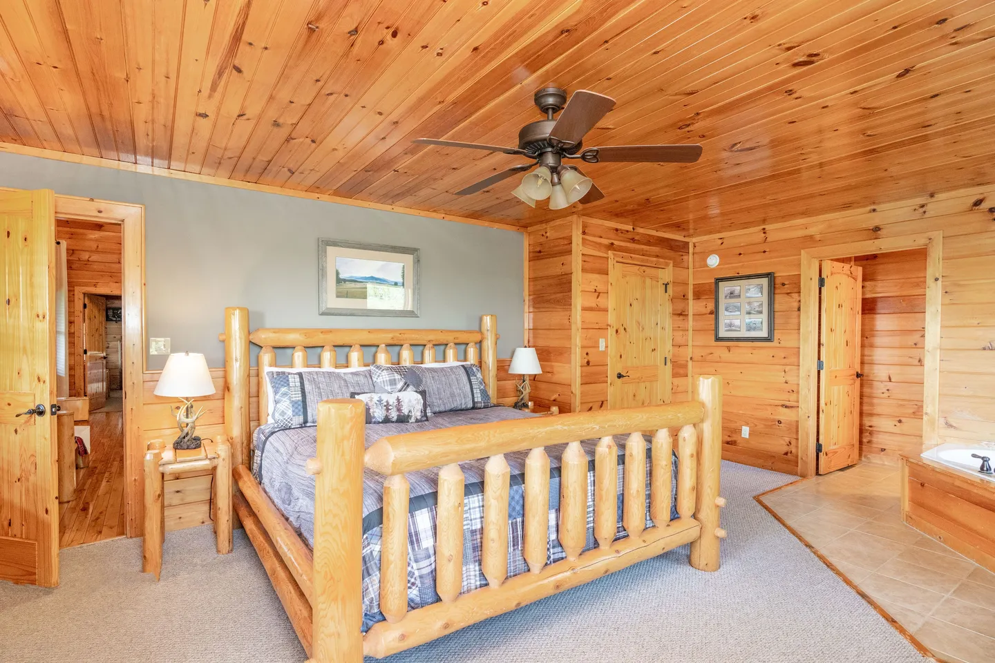 Bedroom with log bed frame, wood-paneled walls and ceiling, carpeted floor, ceiling fan, and an in-room jetted tub.