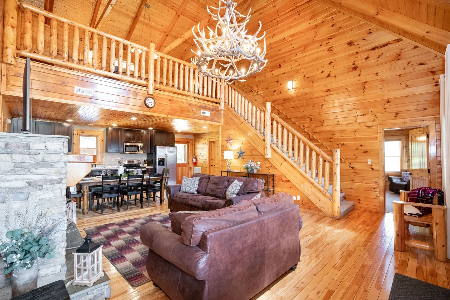Open-concept cabin interior with log-paneled walls, vaulted ceiling with an antler chandelier, living room seating, and a view of the dining area and kitchen.