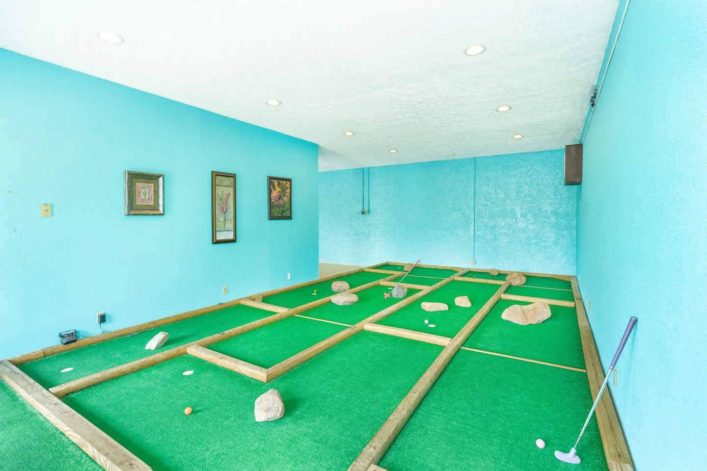 Indoor miniature golf course room with turquoise walls, artificial turf, wooden borders, and rocks as obstacles.