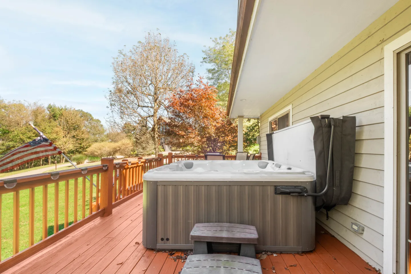 A hot tub with an open cover sitting on a wooden deck next to a house overlooking a grassy yard with trees.