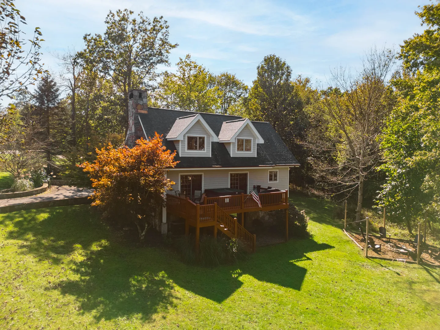 Aerial view of a two-story home with dormers, a stone chimney, and an elevated back deck overlooking a grassy yard bordered by trees.