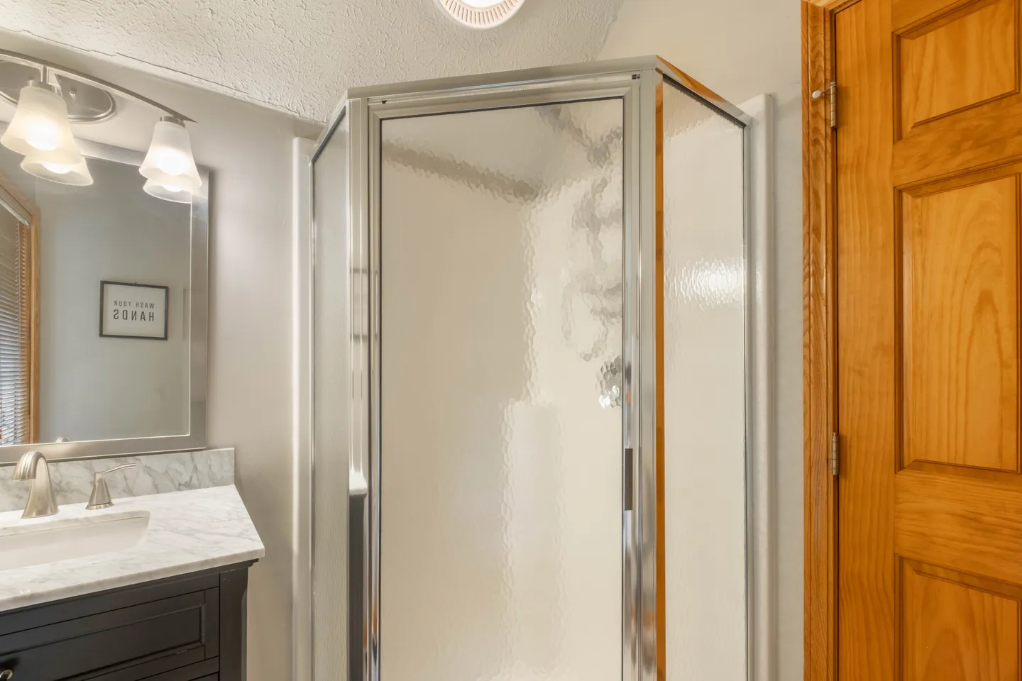 Bathroom corner showing a textured glass shower enclosure adjacent to a dark vanity with marble countertop and a wooden door.