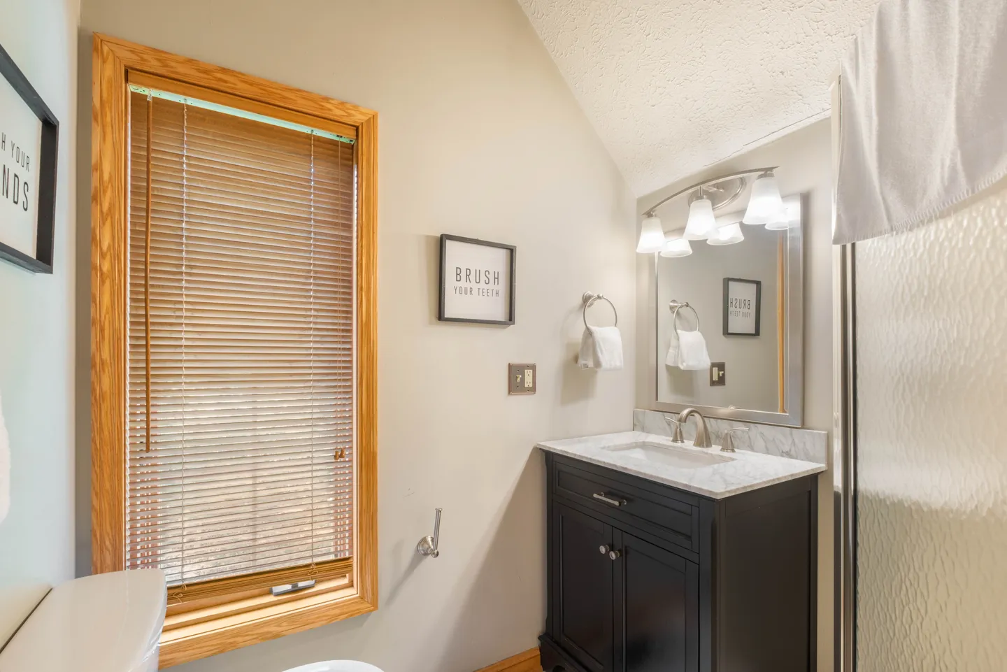 Bathroom with a dark vanity, marble countertop, framed mirror, and a window with wood trim and blinds.