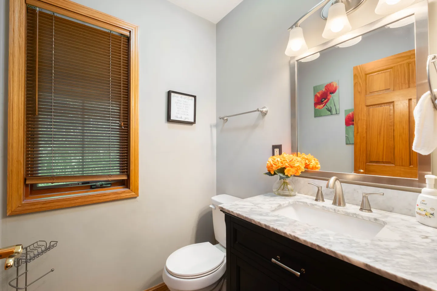 Bathroom with a dark vanity, marble-patterned countertop, mirror with overhead light fixture, toilet, and a window with blinds.