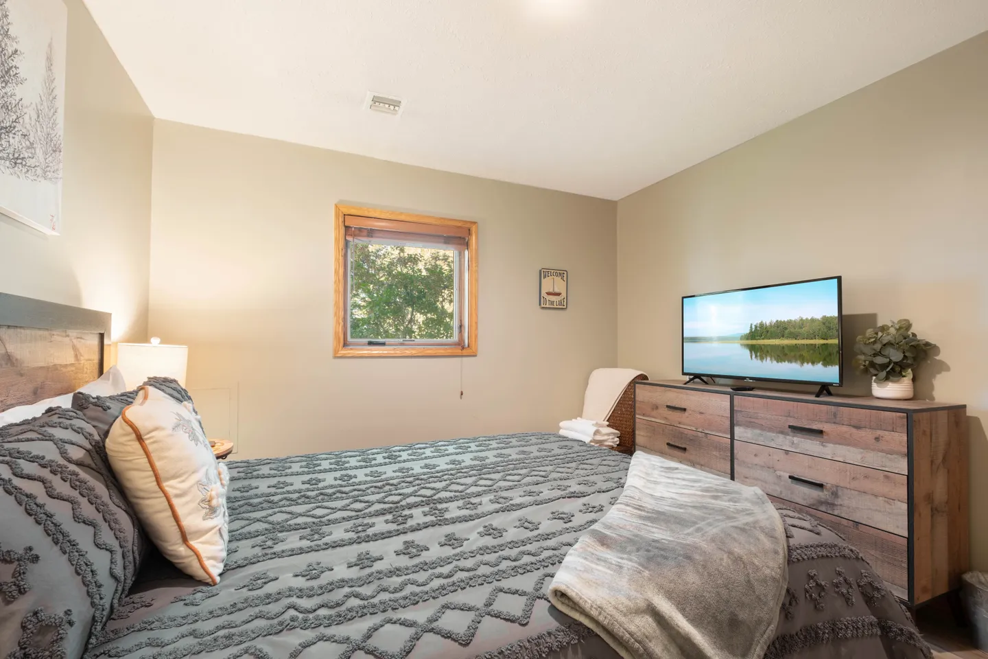 Bedroom with a patterned bedspread, wood-framed window, wooden dresser with a flat-screen TV, and an accent chair in the corner.