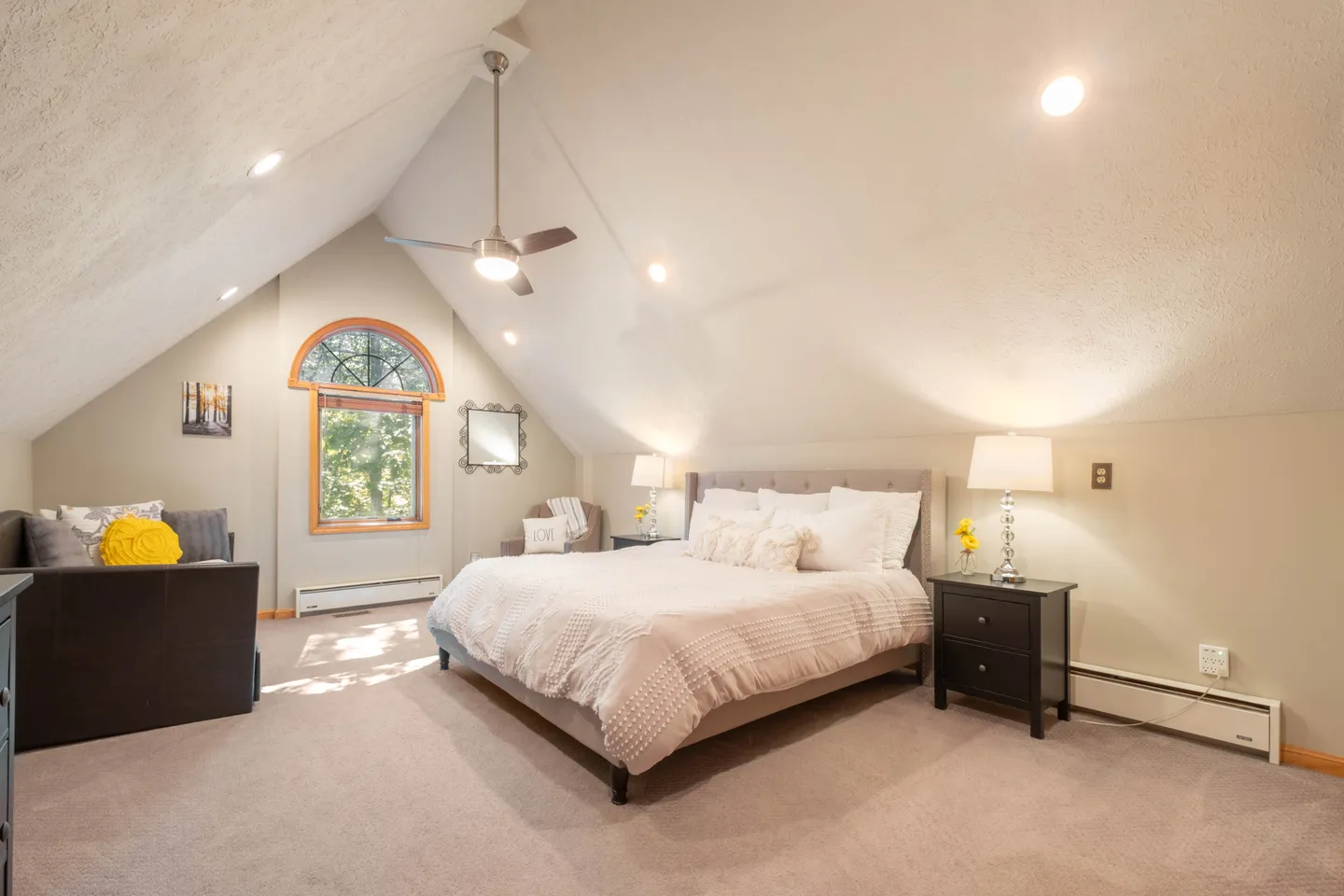 Spacious bedroom with sloped ceilings, carpet flooring, a large bed flanked by nightstands, an armchair, and a sofa under a vaulted roofline.